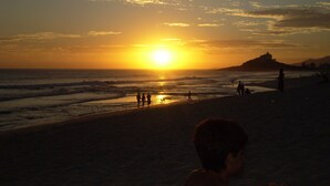 A beach scene at sunset with silhouettes of people and a castle-like structure in the distance. - Furnished apartment on Itaúna beach, a popular spot for international surfing. (Saquarema)