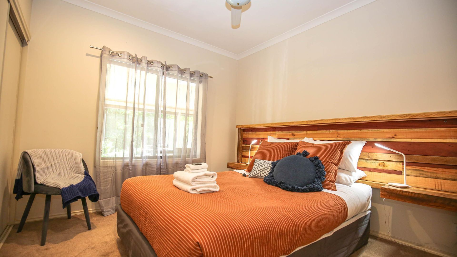 A bedroom with a wooden headboard, a bed with an orange bedspread, a chair with a blanket, and a window with sheer curtains.