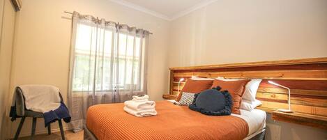 A bedroom with a wooden headboard, a bed with an orange bedspread, a chair with a blanket, and a window with sheer curtains.