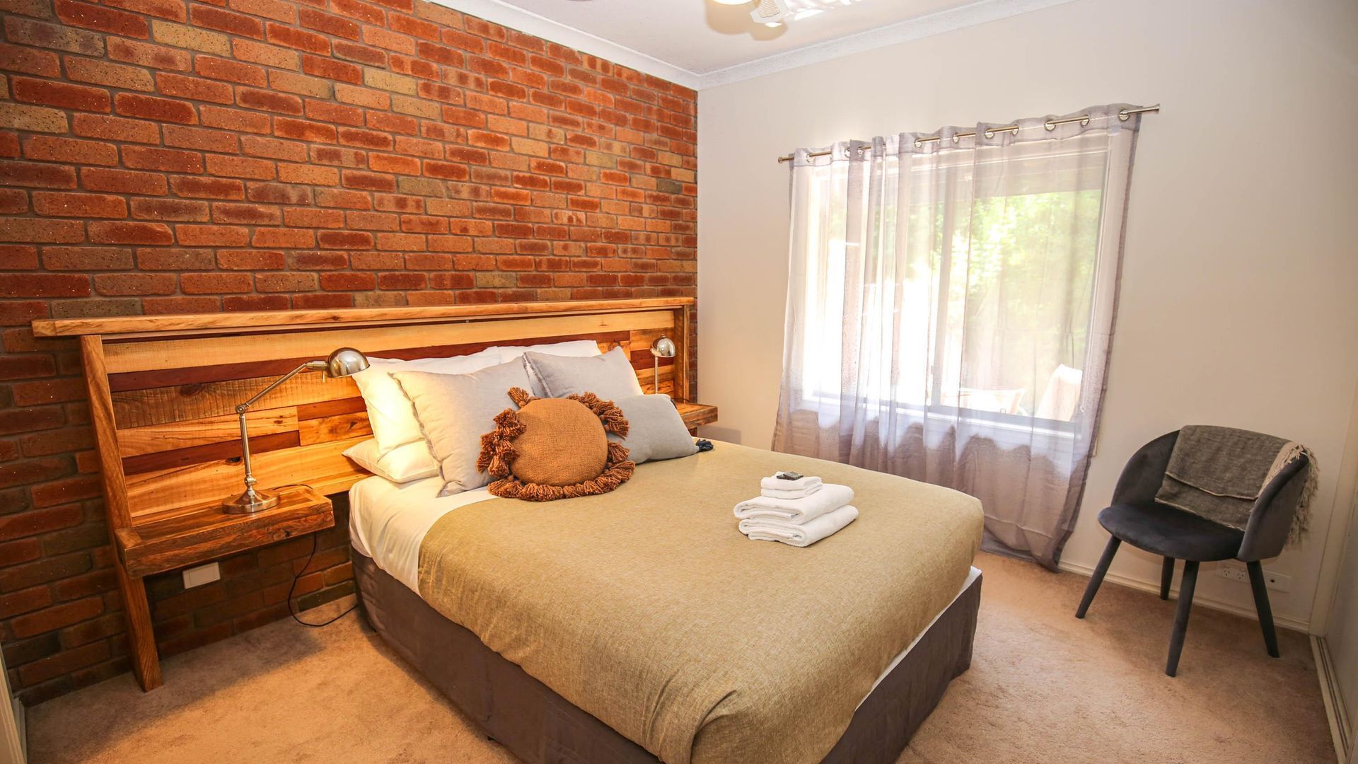 Une chambre avec un mur en briques, une tête de lit en bois, un lit recouvert d’une couverture beige, des oreillers et une lampe de chevet. On y trouve également une chaise et une fenêtre ornée de rideaux légers.
