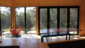 A room with a wooden floor, a table, two chairs, and large windows with a forest view. - BAY OF FIRES ECO HUT Binalong Bay - Off Grid Experience (Binalong Bay)