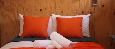Une tête de lit en bois, agrémentée de deux coussins orange et d’un coussin blanc. Des serviettes roulées sont posées sur le lit.