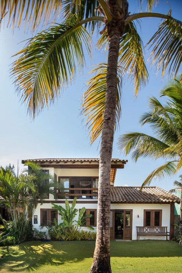 A two-story house with a tiled roof, surrounded by lush greenery and palm trees. - Vila do Outeiro em frente a praça da igreja (Porto Seguro)