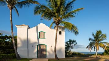 Igreja branca com porta de madeira e molduras de janelas verdes, cercada por palmeiras e um céu limpo.