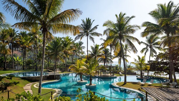 A resort pool area with multiple circular pools, surrounded by palm trees and a wooden deck with lounge chairs. - Vila do Outeiro em frente a praça da igreja (Porto Seguro)