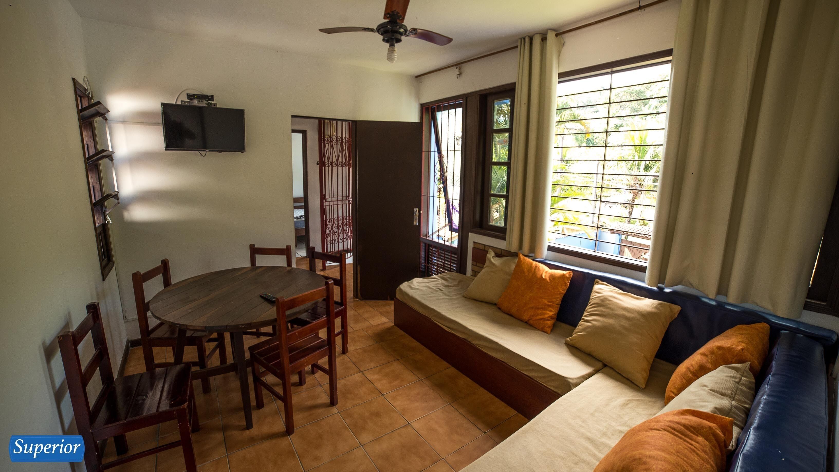 A room with a dining table and chairs, a sofa with cushions, a ceiling fan, and a TV mounted on the wall.