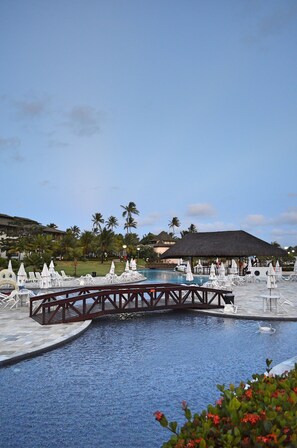 A pool area with lounge chairs, a bridge, and a thatched-roof structure. - Luxury Bungalow at Beach Class Resort Muro Alto FOR RENT (Porto de Galinhas)