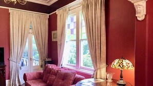 Ett rum med en röd vägg, en rosa soffa, ett runt träbord med en lampa och ett fönster med gardiner.