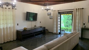 Een woonkamer met een grote bank, een aan de muur bevestigde televisie, een houten plafond en uitzicht op groen buiten.
