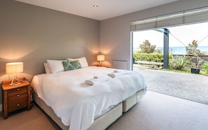 Quarto moderno com uma cama grande, abajures e vista para um jardim e mar através de uma porta de vidro deslizante.
