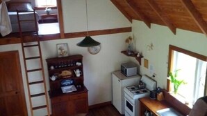A compact kitchen with a stove, microwave, and small refrigerator. A wooden ladder and shelves with kitchenware are also visible. - Ruru Cottage - cosy native forest hideaway (Pakawau Beach)