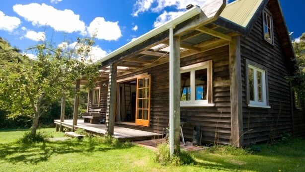 A rustic wooden cabin with a porch, large windows, and a sloped roof. - Ruru Cottage - cosy native forest hideaway (Pakawau Beach)
