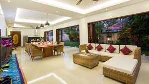 A spacious living area with a dining table, wicker furniture, and a large window overlooking a garden. - La Bali 4 Bedrooms Villa Sanur (Denpasar Selatan)