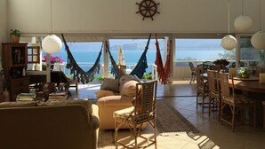 Uma sala de estar espaçosa com vista para o mar, uma rede, uma mesa de jantar com cadeiras e uma estante de livros.