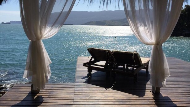 Um deck de madeira com um banco, com vista para a água, emoldurado por cortinas brancas.