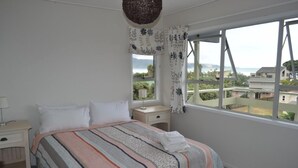 Un dormitorio con un ventanal grande, una cama con manta a rayas, una mesita de noche con lámpara y vistas al exterior con zonas verdes y edificios.
