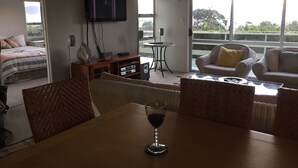 Amplia sala de estar con balcón, televisor, mesa de comedor con una copa de vino y un cómodo espacio para sentarse.
