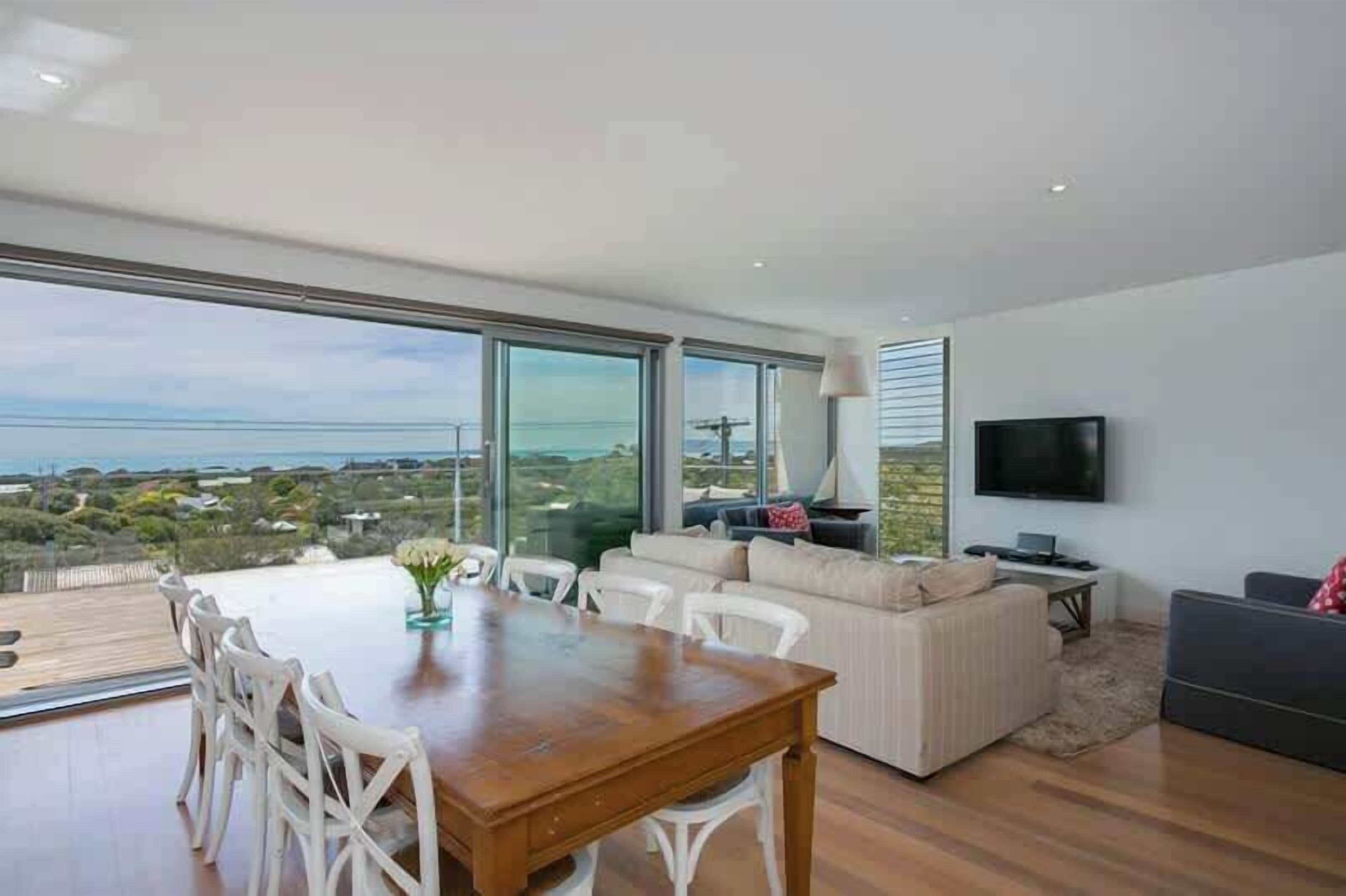 A modern living room with a dining area, large windows, and a flat-screen TV.