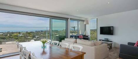 A modern living room with a dining area, large windows, and a flat-screen TV.