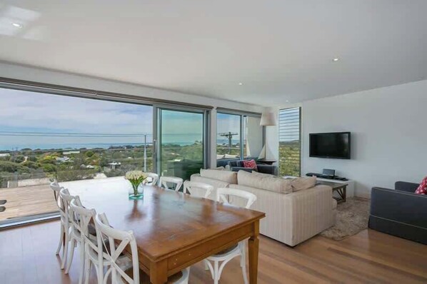 A modern living room with a dining area, large windows, and a flat-screen TV.