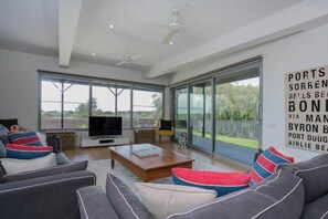 A modern living room with a flat-screen TV, grey sofas, and a large window offering a view of the outdoors.