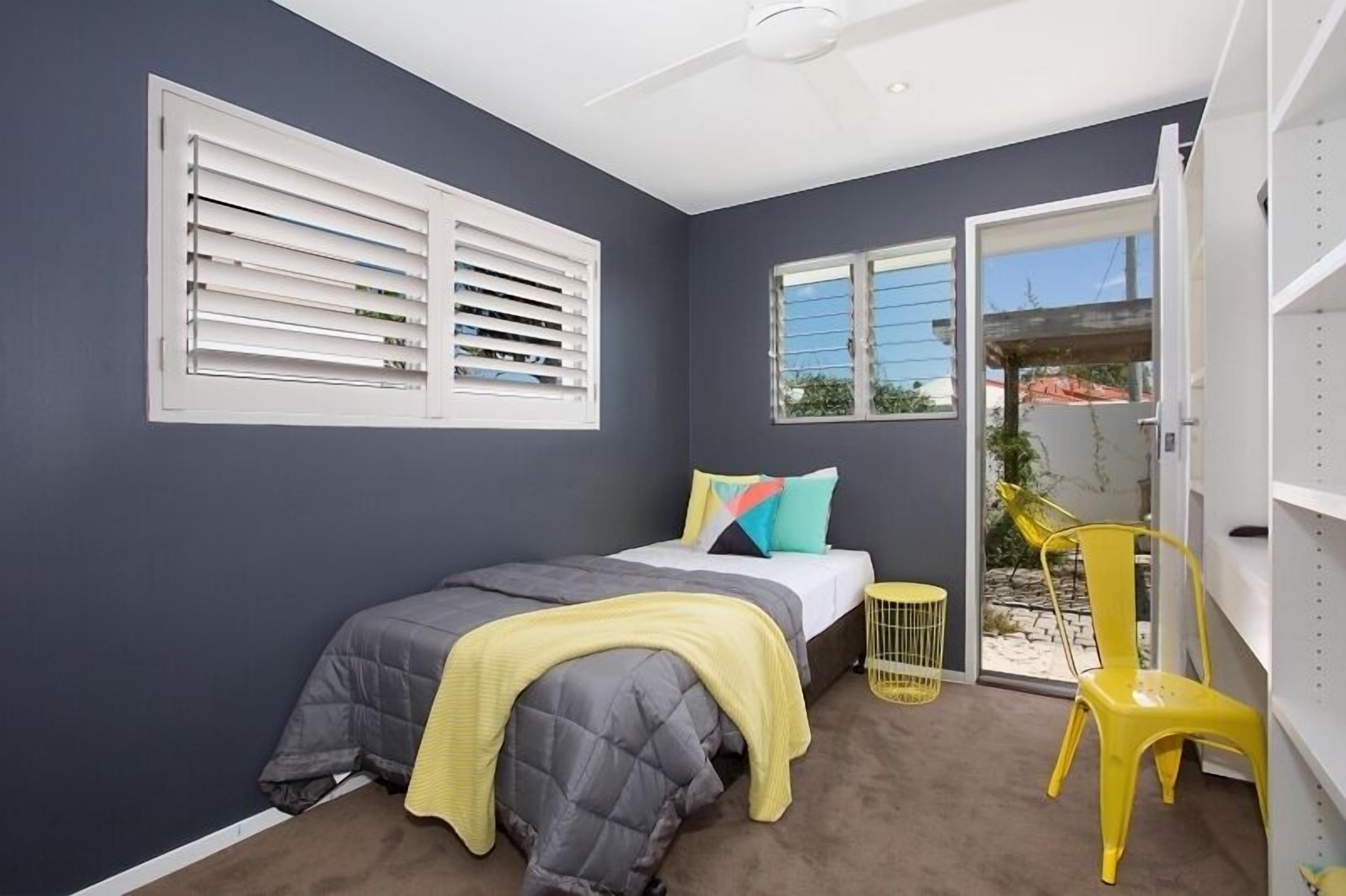 Ein Schlafzimmer mit einem Bett, einem Deckenventilator, einem Fenster mit Jalousien, einem Stuhl und einem kleinen Tisch.