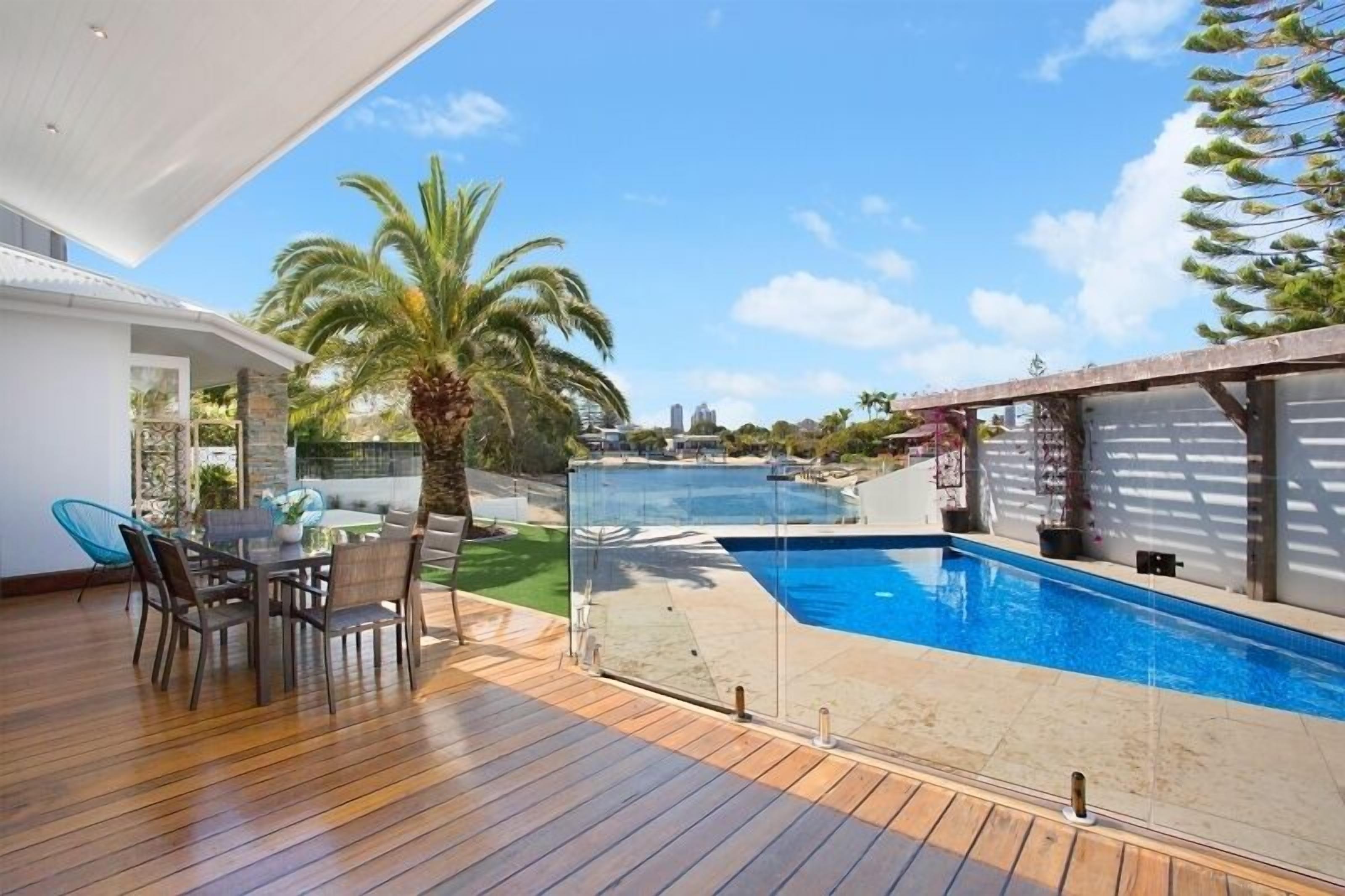 A modern outdoor pool area with a wooden deck, a dining table with chairs, and a view of the ocean.