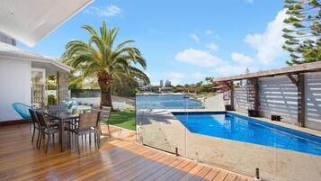 A modern outdoor pool area with a wooden deck, a dining table with chairs, and a view of the ocean.