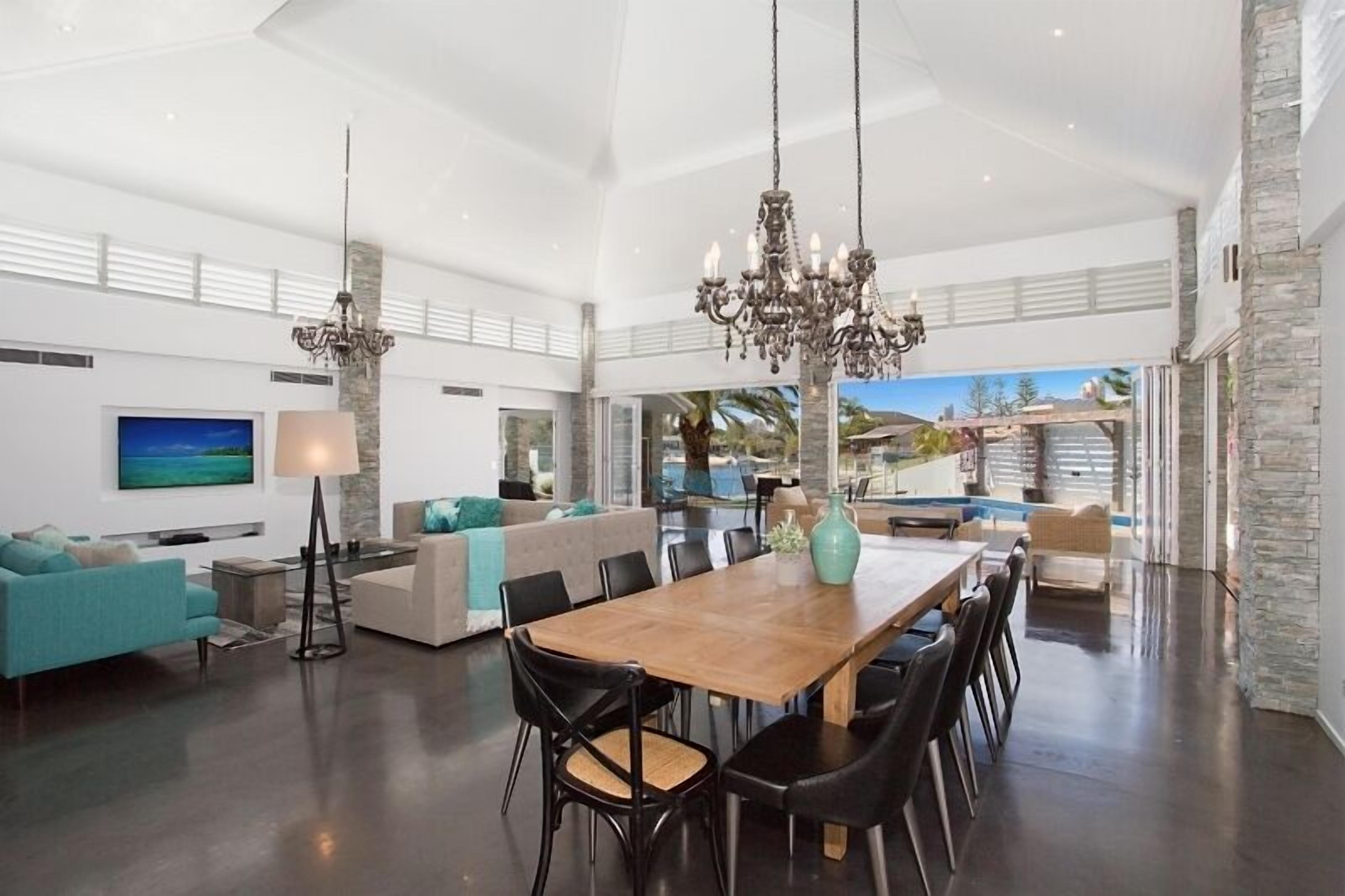 A spacious living area with a large wooden dining table, black chairs, and a chandelier. There is a sofa and a TV mounted on the wall.