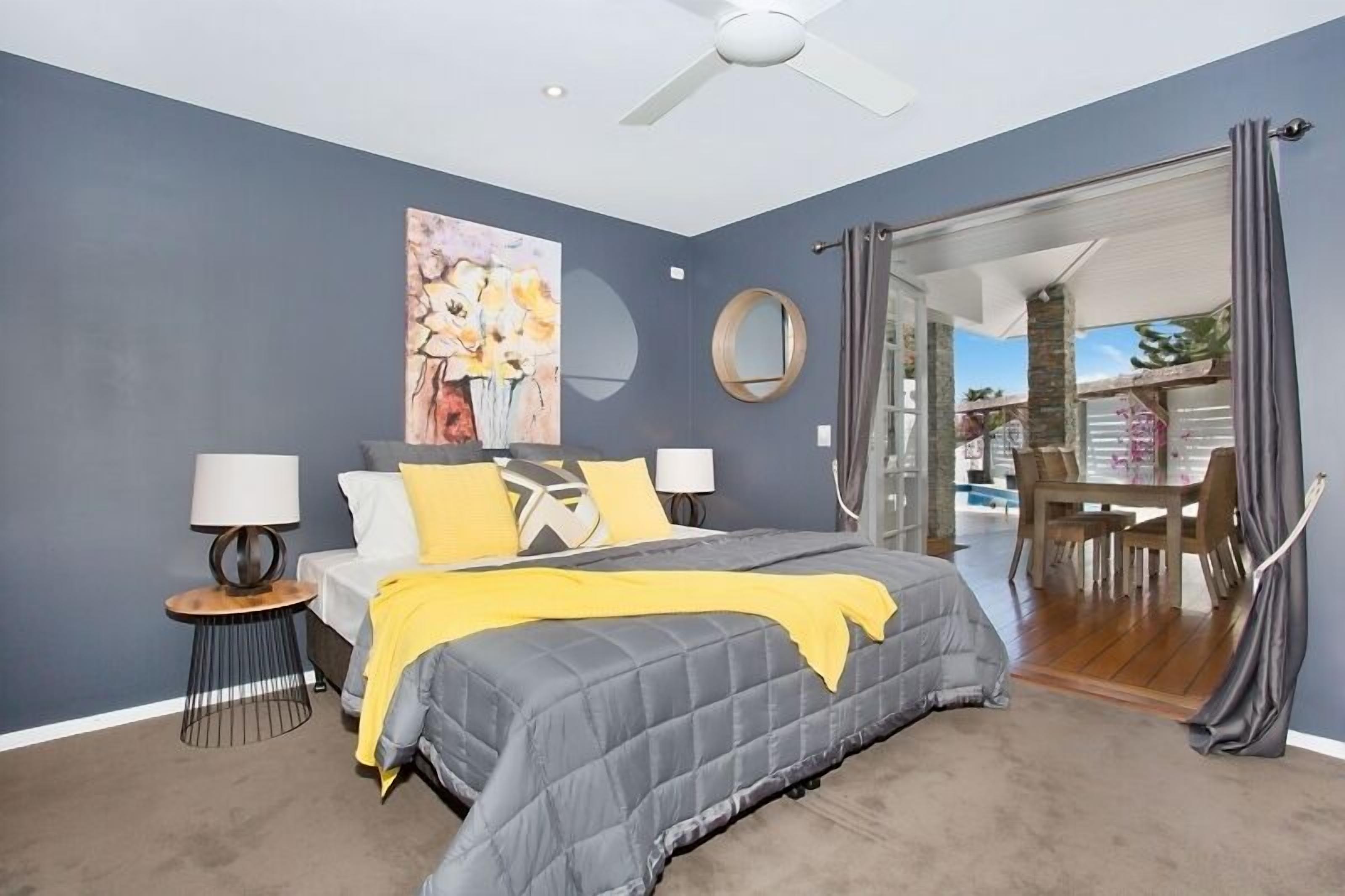A bedroom with a bed, bedside lamps, a ceiling fan, a painting, and a view of an outdoor area with a table and chairs.