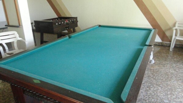 A pool table with a green felt surface, a foosball table, and a white chair in a room with sloped ceilings.