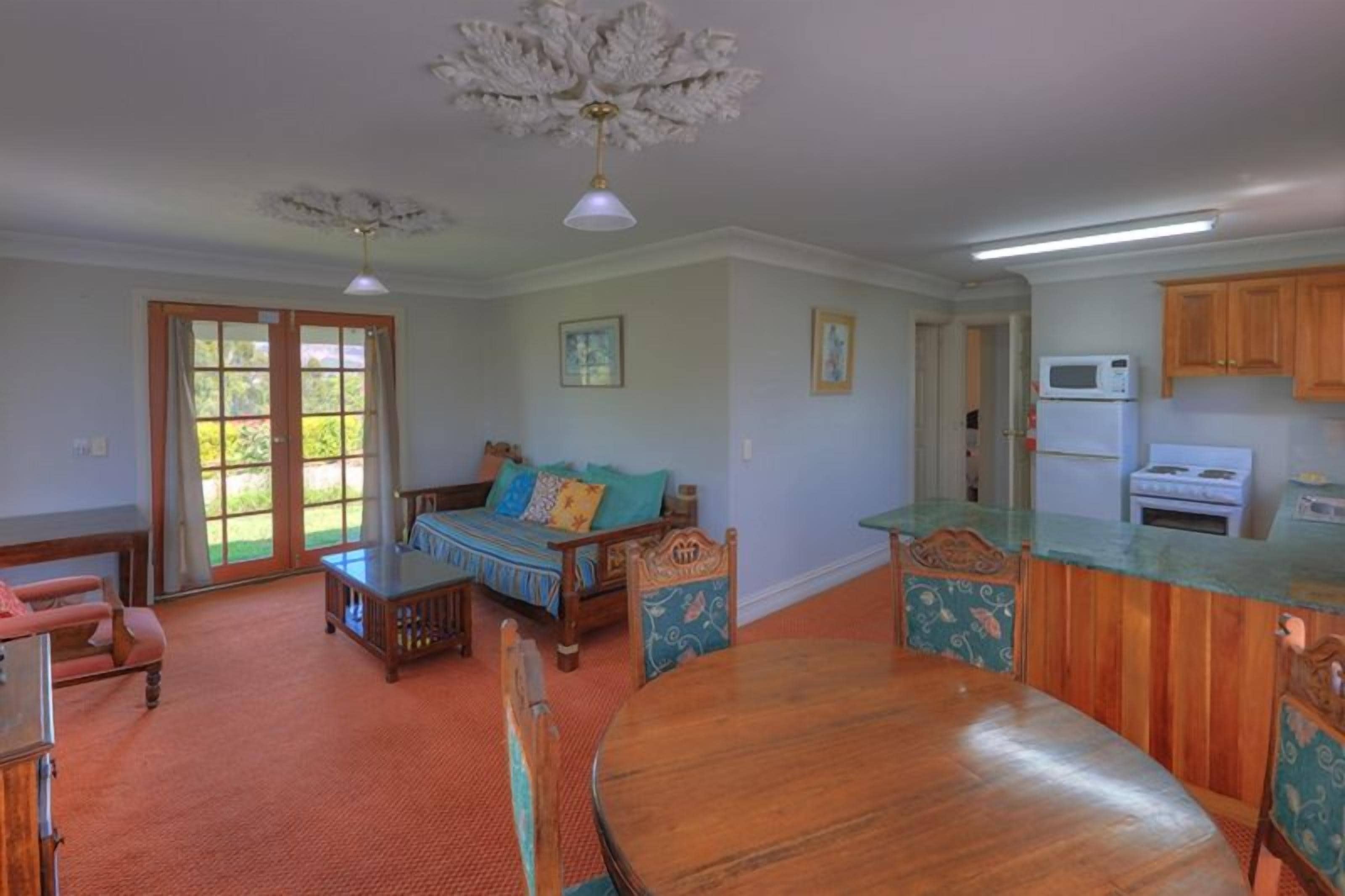 A spacious living room with a wooden dining table, a sofa with patterned cushions, and a kitchen area with wooden cabinets and a microwave.