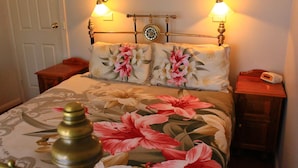 A bedroom with a floral bedspread, two bedside tables, a nightstand, a bedside lamp, and a wall-mounted clock. - Hatcher's Manor Richmond - Farmhouse (Richmond TAS)