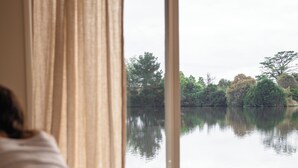 Una persona sdraiata su un letto con vista su un lago calmo e alberi, visibili attraverso una finestra con tende.