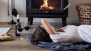 Una stanza con una stufa a legna in legno nero, una persona sdraiata sul pavimento, una bottiglia di vino e dei bicchieri.
