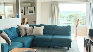 A teal sectional sofa with multiple cushions, a wooden coffee table, a framed picture on the wall, and a view of greenery through a window. - Werri Escape - walk to the beach (Gerringong)