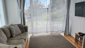 A living room with a sofa, a TV, a small table, and a view of a residential street through a large window. - Werri Escape - walk to the beach (Gerringong)