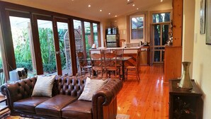 A spacious living area with a leather sofa, wooden dining table, and chairs. A kitchen with a refrigerator and microwave is visible in the background. - Bishops Garden Spa Suite (North Adelaide)