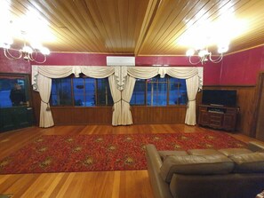 Ein Zimmer mit Holzboden, einem großen Fenster mit Vorhängen, einem Fernseher und einem Ledersofa.
