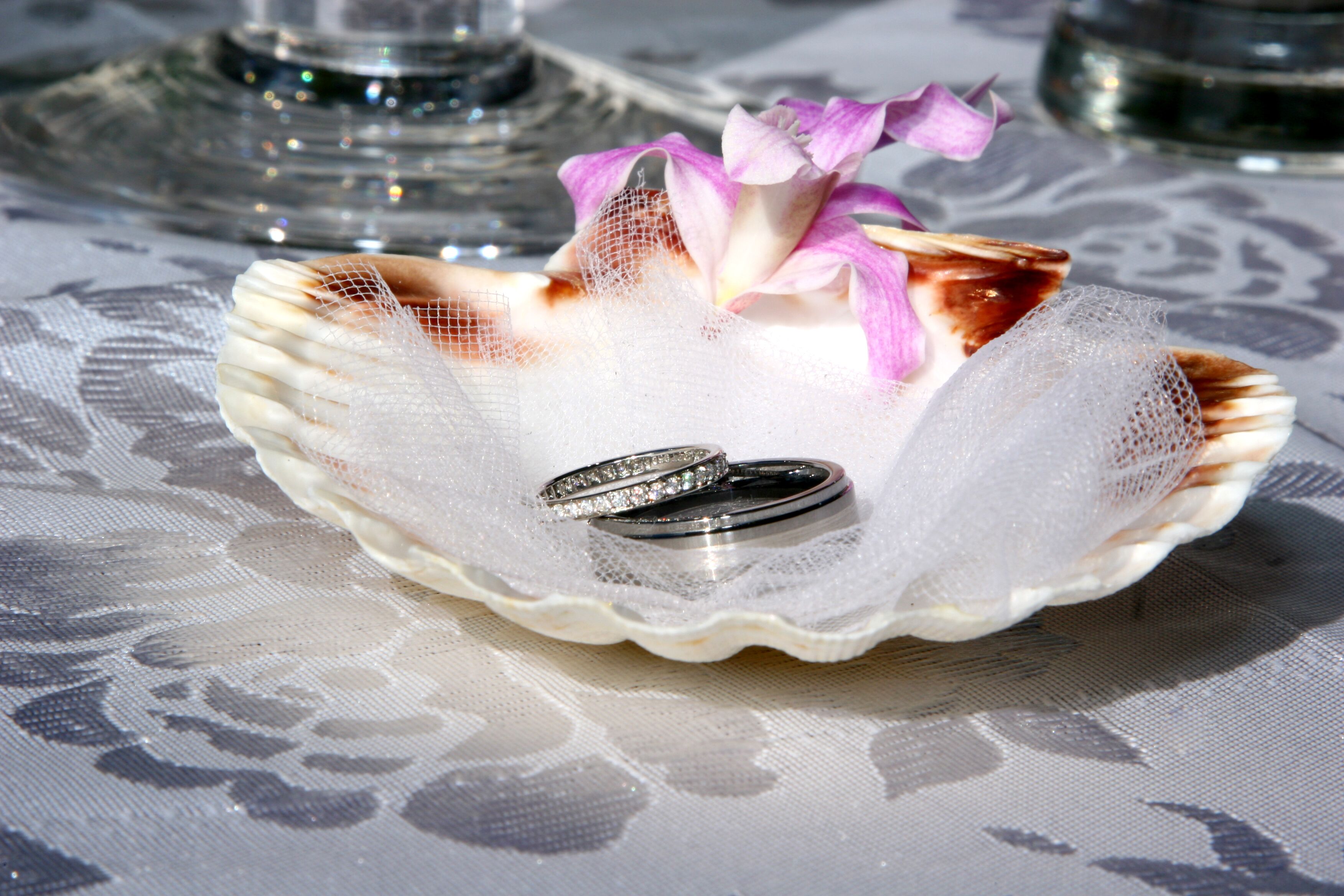 Una concha decorativa con dos anillos y una flor sobre un mantel de encaje.