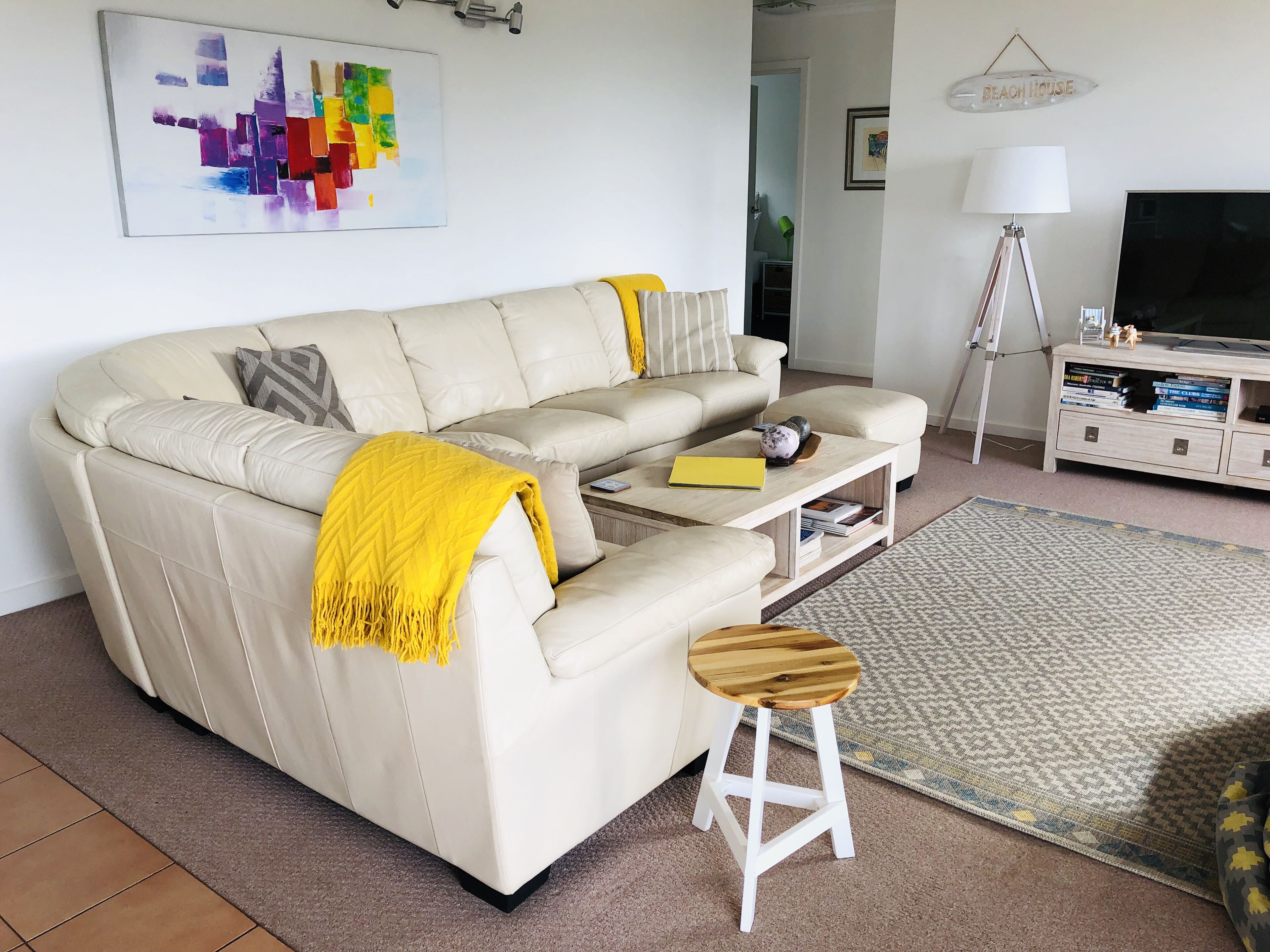 A living room with a beige sofa, a yellow throw blanket, a wooden stool, a coffee table, a lamp, a TV, and a bookshelf.