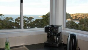 En kaffemaskine på en køkkenbordplade med udsigt til havet og træer gennem et vindue.