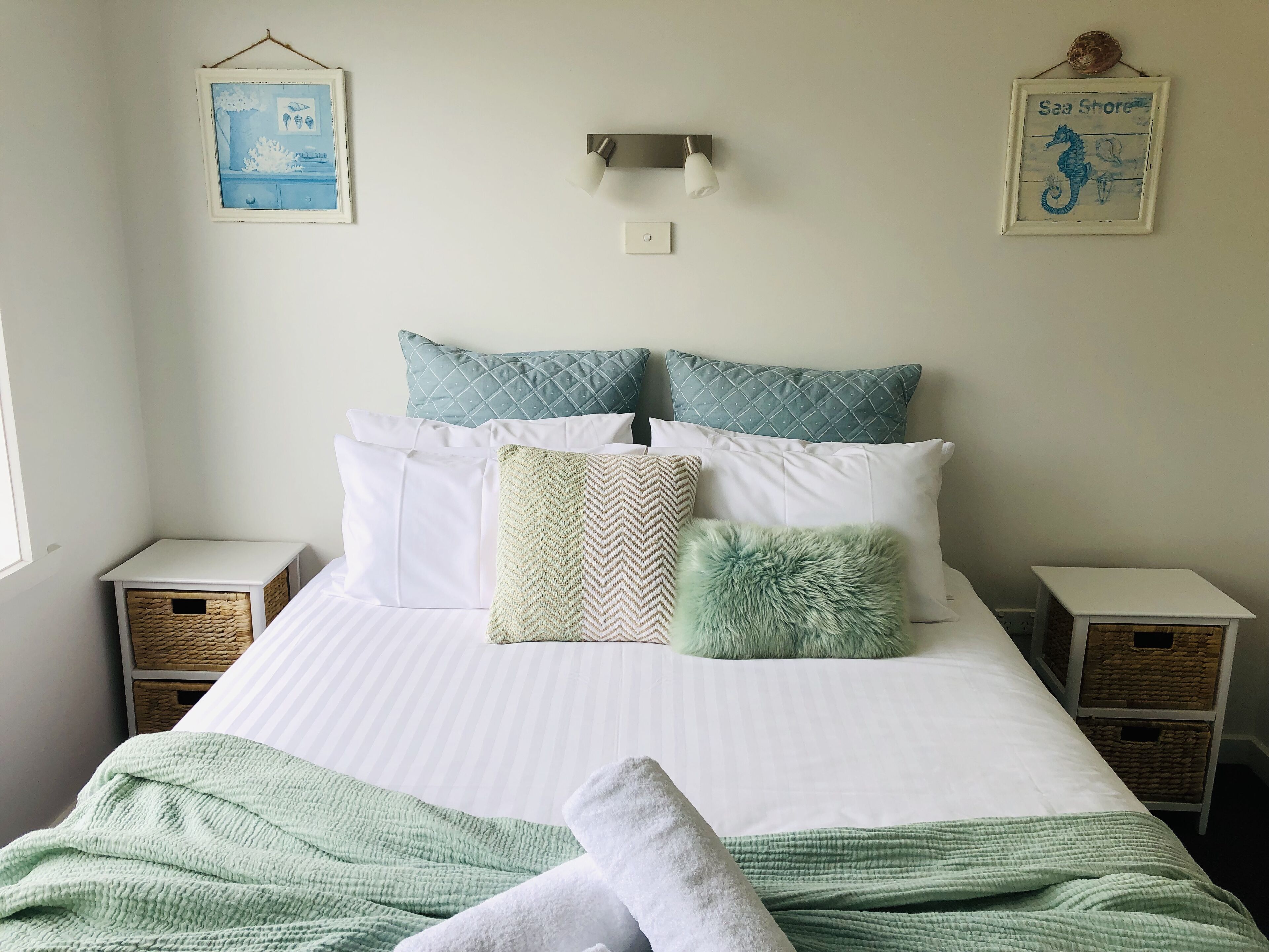 A neatly made bed with a striped pillowcase, a green blanket, and a white towel. There are two nightstands with wicker baskets, a wall-mounted light, and two framed pictures.