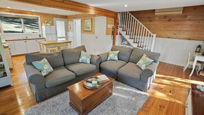 A cozy living room with a gray sectional sofa, a wooden coffee table, and a view of the kitchen and staircase.