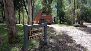 A wooden sign with the text "Kookas Cottage" in a forested area with a cabin in the background.