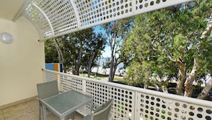 Un balcon agrémenté d’une table et de chaises, entouré d’une balustrade blanche ajourée, donnant sur une plage et des arbres.