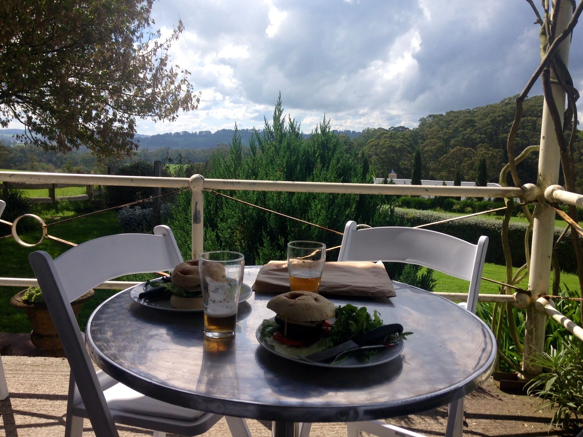 A round metal table with two white chairs, a glass of beer, and a burger on a plate.