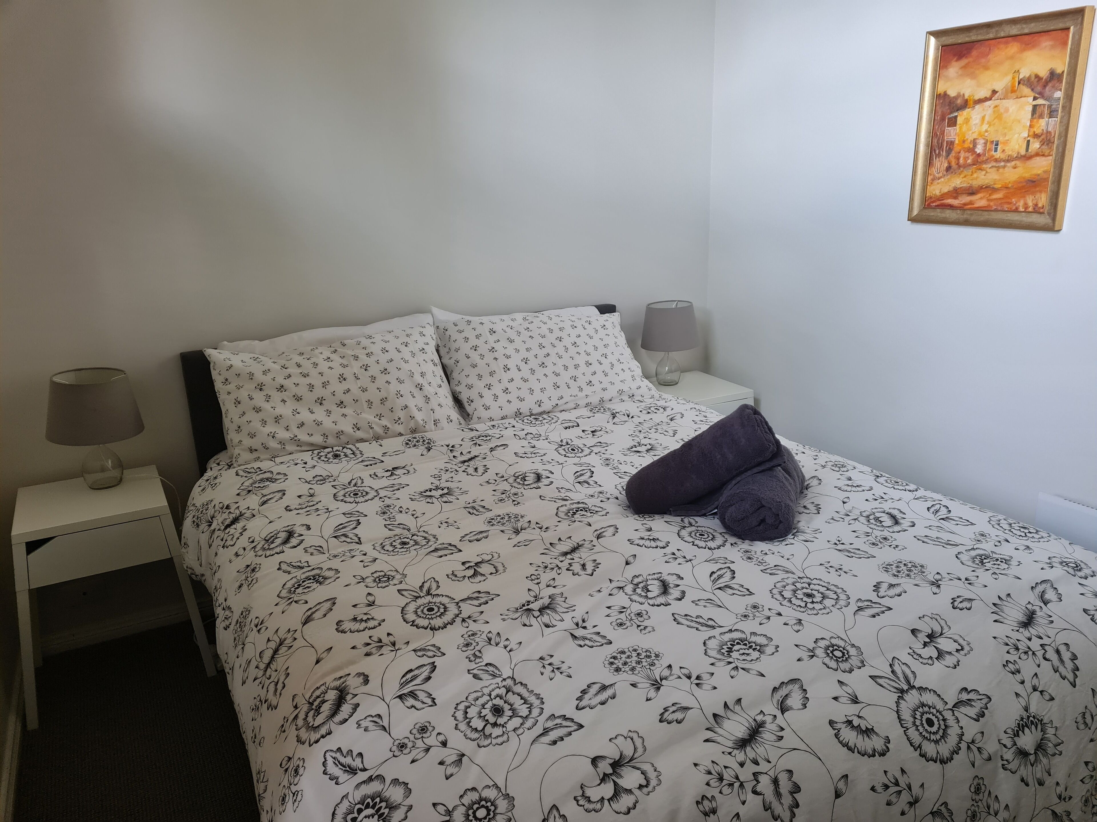 Un dormitorio con una colcha estampada con motivos florales, dos almohadas, una mesita de noche con una lámpara y un cuadro en la pared.