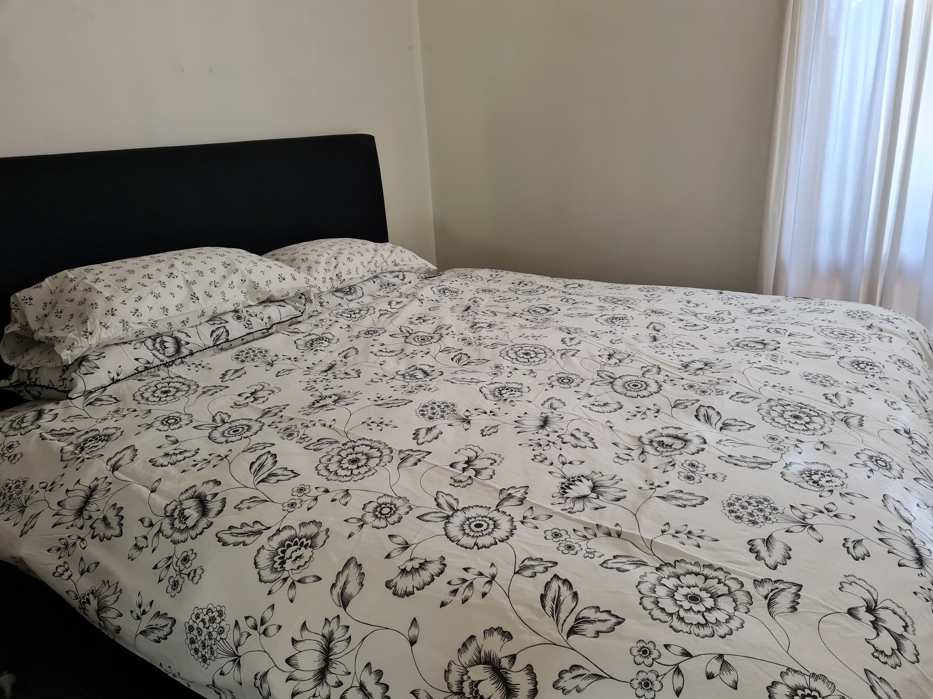 Un lit avec une couvre-lit et des oreillers à motifs floraux, une tête de lit sombre et une fenêtre avec des rideaux.