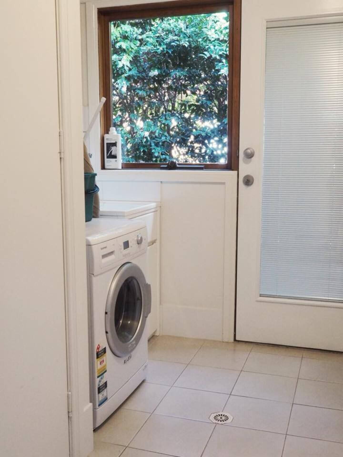Ein kleiner Waschraum mit einer Waschmaschine mit Frontlader, einem Fenster mit Blick auf Bäume und einer Tür mit Jalousie.